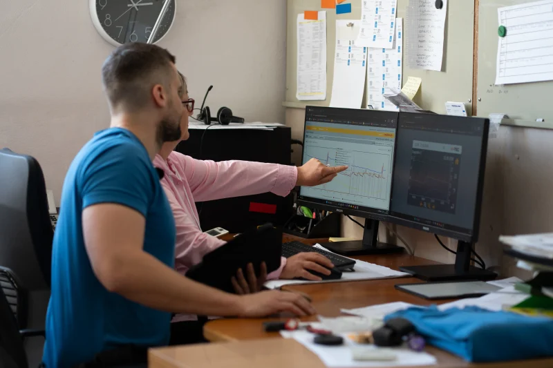Zwei Personen besprechen technische Darstellungen an einem Arbeitsplatz mit zwei Bildschirmen.