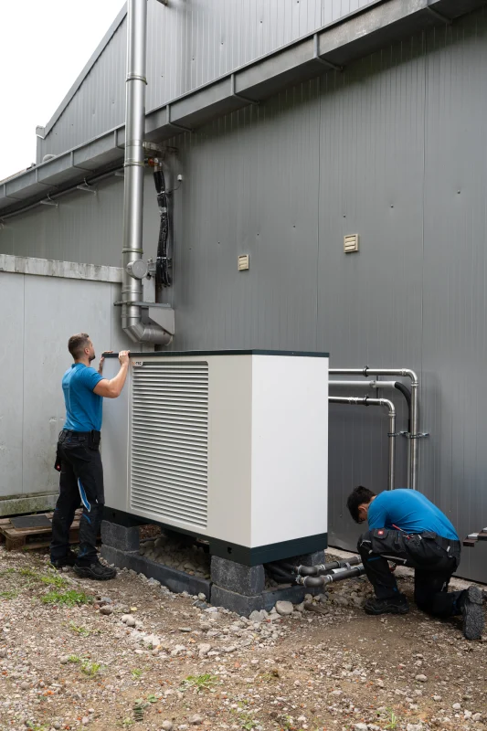 Zwei Monteure installieren eine weiße M-TEC-Außenwärmepumpe vor einer grauen Fassade.