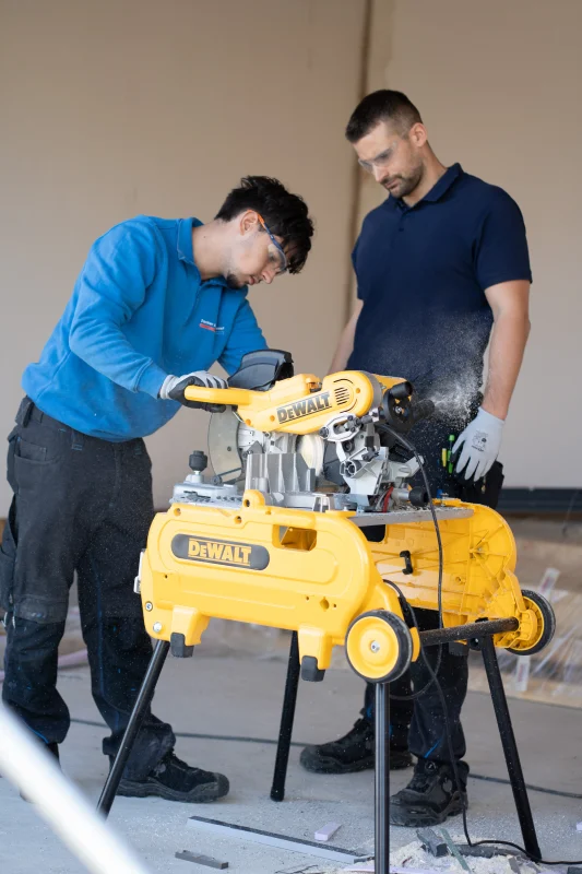 Zwei Mitarbeiter arbeiten mit einer gelben DeWalt-Kappsäge an einem mobilen Arbeitstisch im Rohbau.
