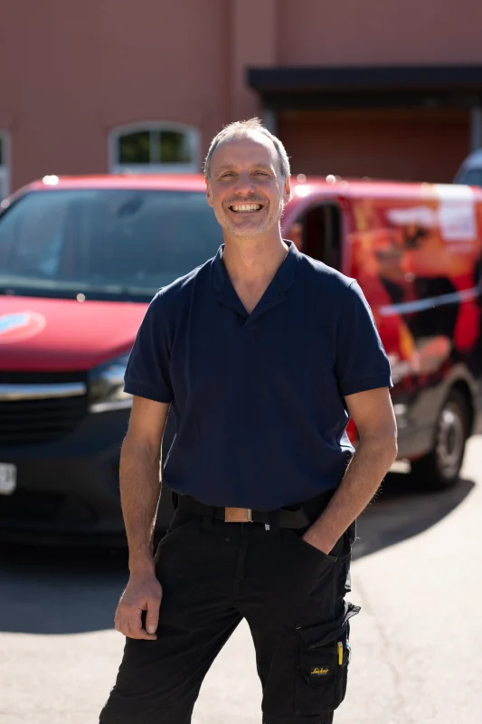 Ein älterer Mann im dunkelblauen Poloshirt steht lächelnd vor roten Firmenfahrzeugen.