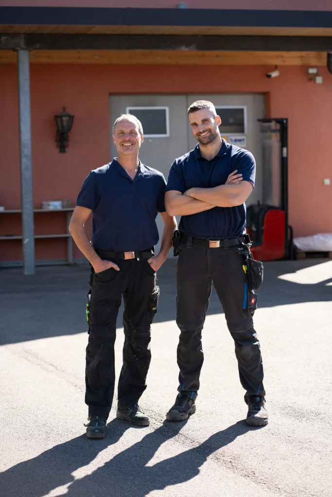 Zwei Männer in dunkelblauen Poloshirts stehen vor einem rosafarbenen Gebäude.