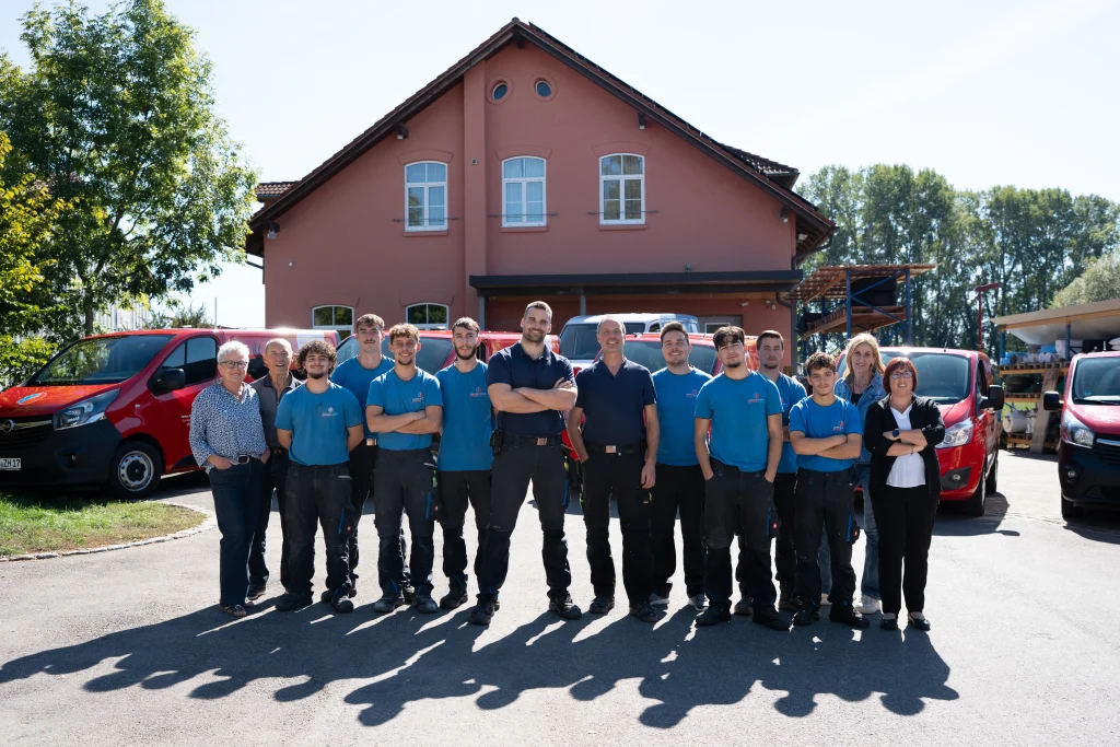 Das Team steht vor dem rosafarbenen Firmengebäude neben roten Firmenfahrzeugen.
