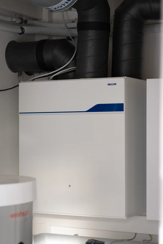 Ein weißes WOLF-Gerät ist an schwarze gedämmte Luftkanäle im Technikraum angeschlossen.