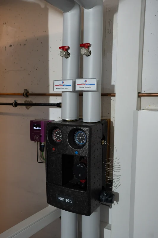 Eine schwarze Pumpengruppe mit zwei runden Anzeigen ist an weiße gedämmte Leitungen mit roten Absperrgriffen angeschlossen.