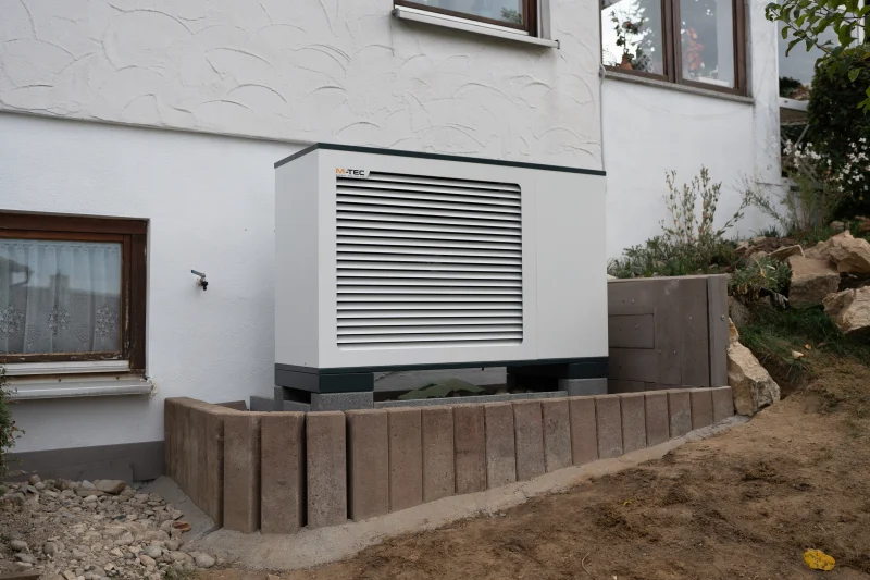 Eine weiße Wärmepumpe mit Lamellengehäuse steht neben einem weißen Wohnhaus an einem bepflanzten Hang mit Holzeinfassung.