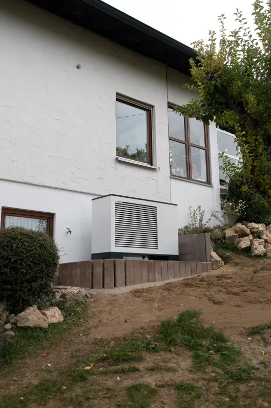 Eine weiße Wärmepumpe mit Lamellengehäuse steht neben einem weißen Wohnhaus an einem bepflanzten Hang mit Holzeinfassung.