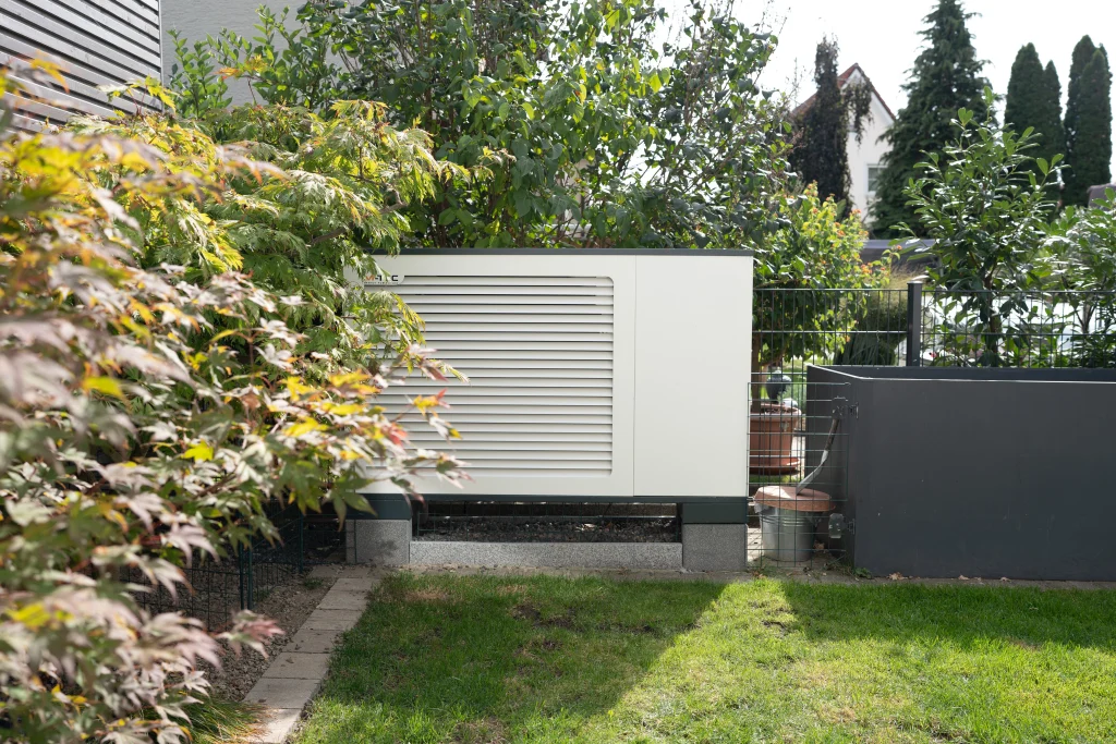 Eine weiße Wärmepumpe mit Lamellengehäuse steht in einem bepflanzten Garten neben grauen Hochbeeten.